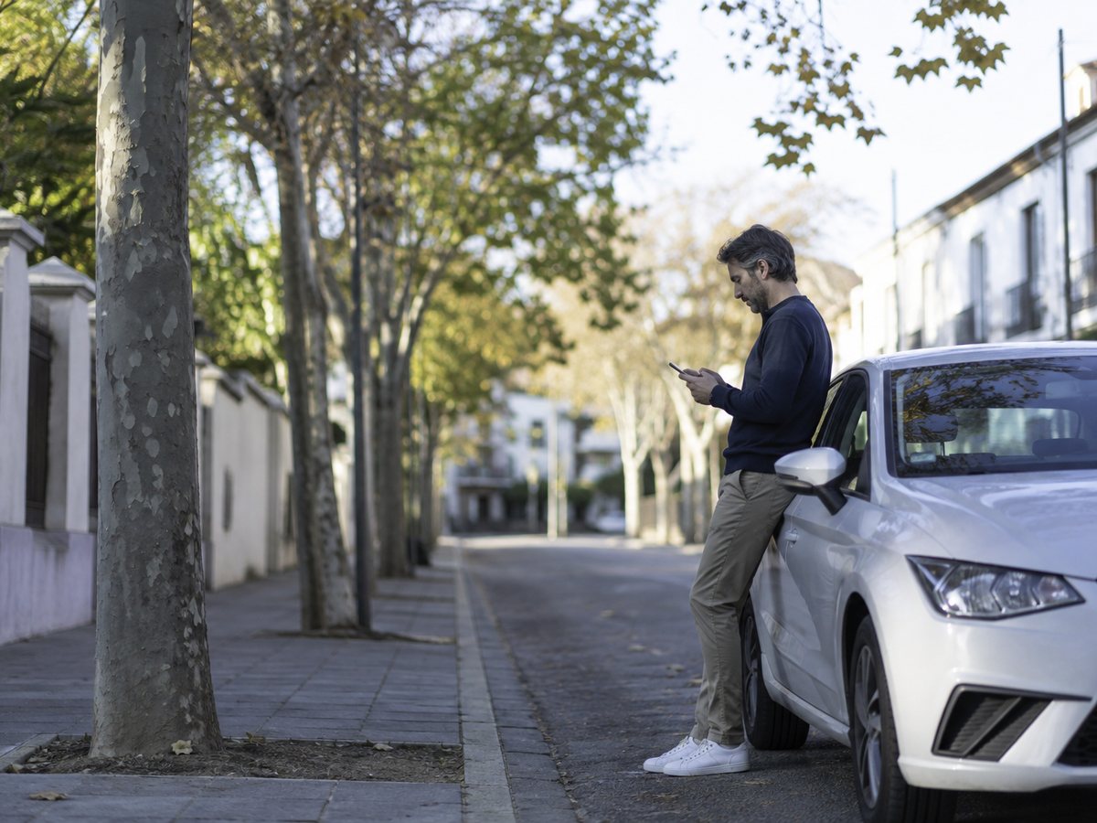 Un conductor consulta el móvil junto a su coche en una calle arbolada